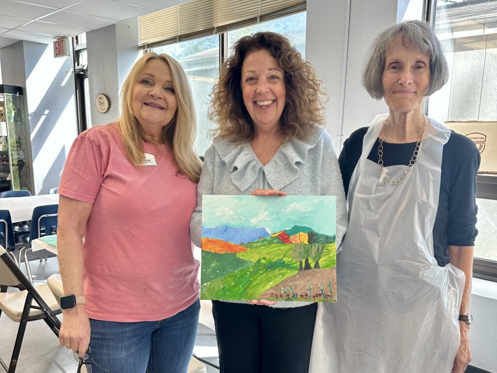 Three women one of whom is holding a landscape painting, created at Massanutten Resort.