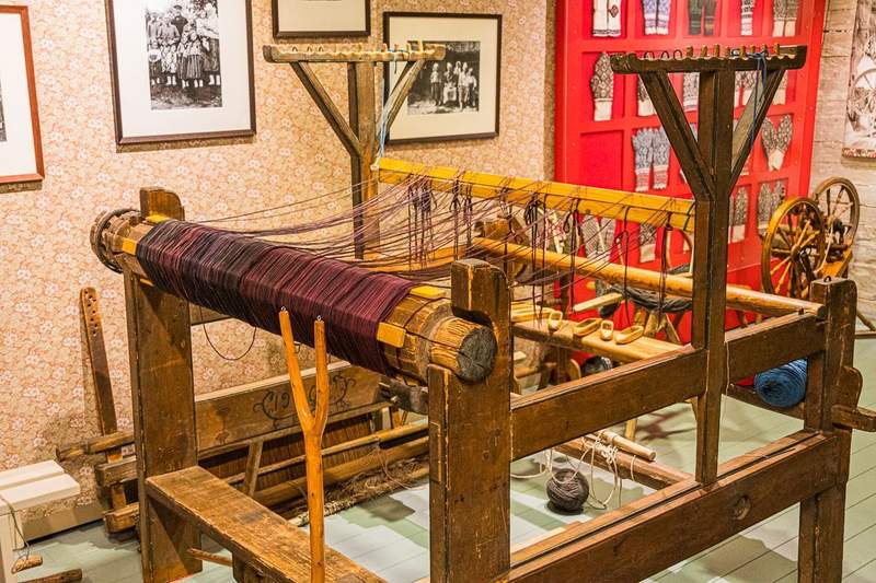 An old wooden frame loom showing the warp rolled onto a large wooden drum and the shuttles, used for creating the colorful fabric, resting on the loom