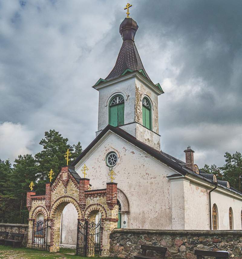 A simple white building with a tower topped by a small onion dome and a gold cross. In front of the church is a stone wall and brick gated entrance to the church grounds.