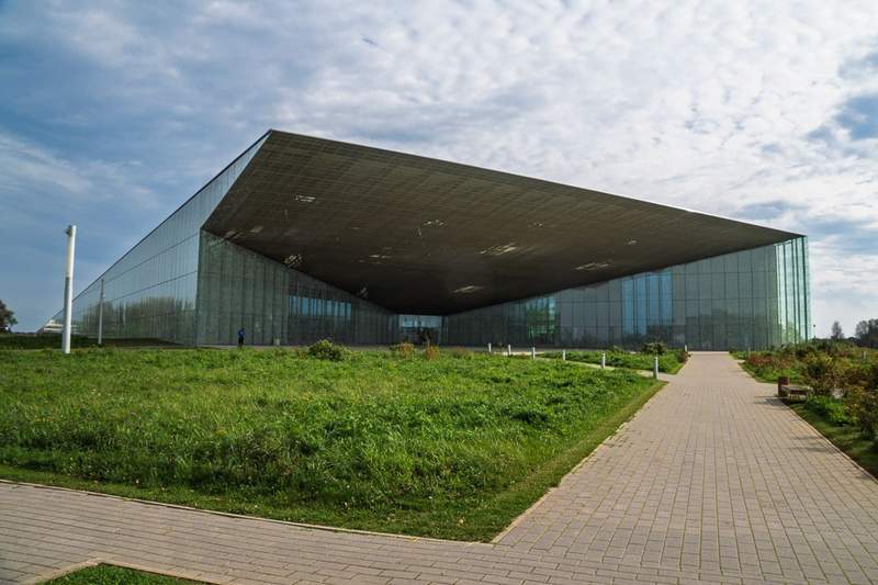 A brick path leading to a wedge shaped building with a deeply recessed front entranceand having the appearance of being entirely constructed of glass panels.