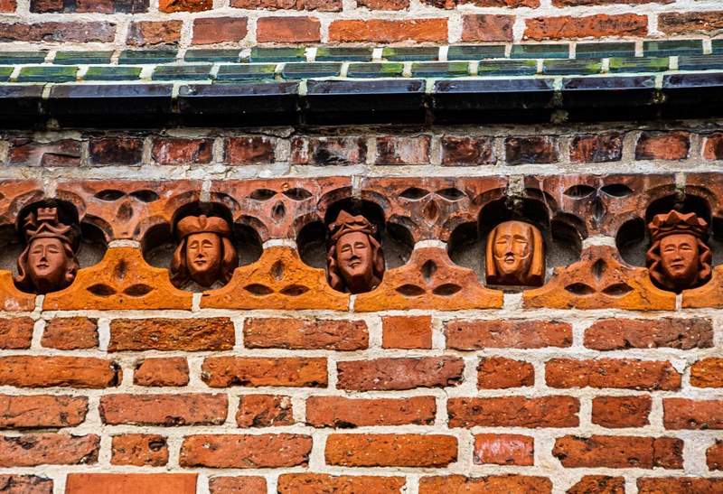 A small section of an exterior red brick wall with five recesses each containing a small terracotta face