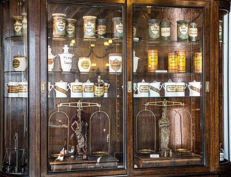 A tall glass fronted  wooden cabinet displaying different shaped ceramic vessels for medicine ingredients and old apothecary scales.