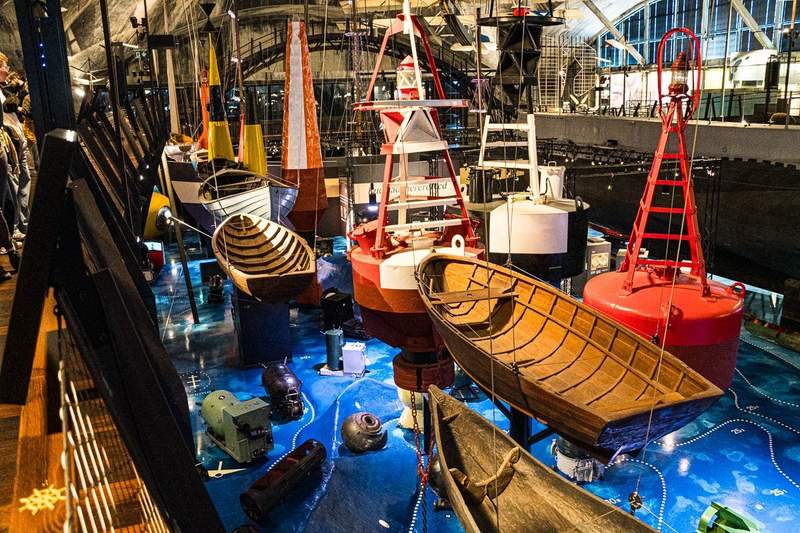 View from an elevated walkway of exhibits that include wooden row boats and navigation buoys hanging from the ceiling and mines and other underwater objects on the floor. 