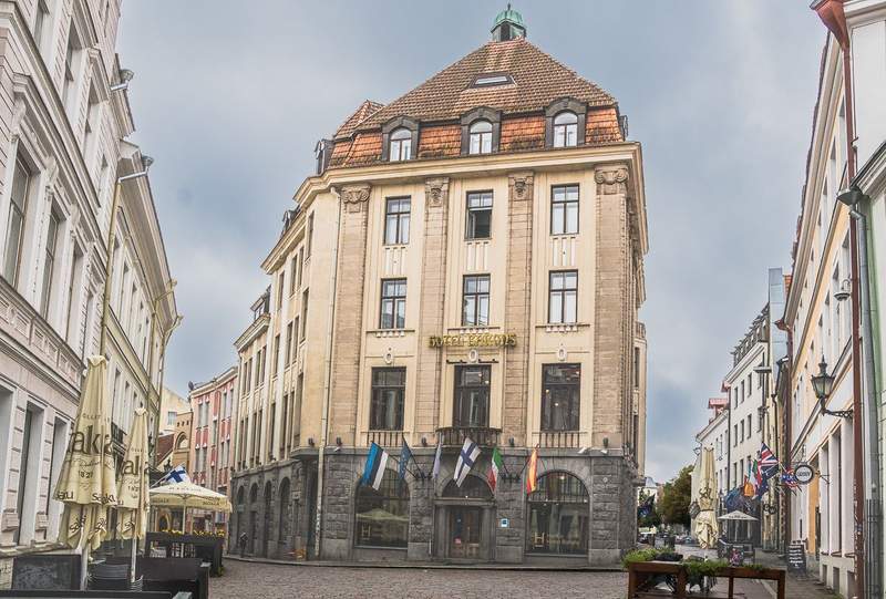 A five storey stone art nouveau building with a red tile roof and cobbled streets on either side of the building. Flags are displayed over the entrance.