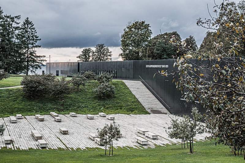 sunken patio of white stone with benches surrounded by apple trees facing a black marble wall part of memorial with names of victims of the Soviet occupation.