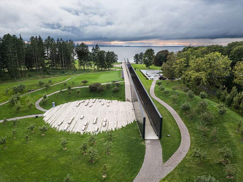 An aerial view of two parallel black walls containing an open passage way leading towards a body of water in a park-like setting that includes a round paved white area with white backless benches.