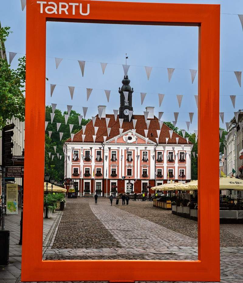 Tartu city hall and main square framed by an orange vertical rectangle with the word Tartu in  white lettering in the top left  corner.