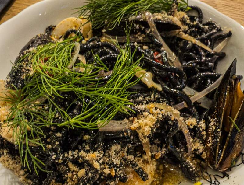 Dill sprigs and grated cheese  scattered on top of black spaghetti and mussels in a white bowl.