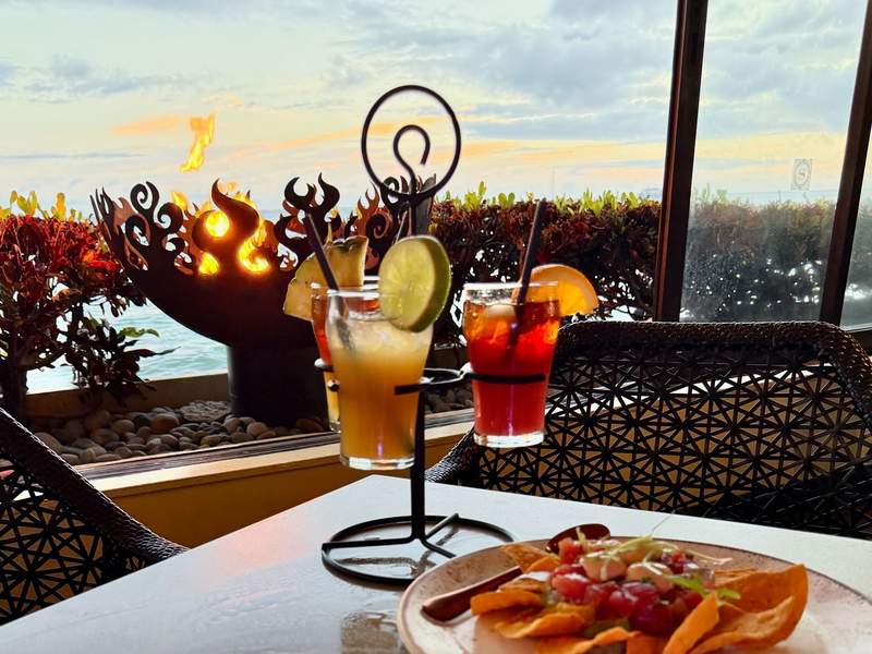 Three colorful mai tais served in a funky metal holder with a firepit and the ocean in the background.