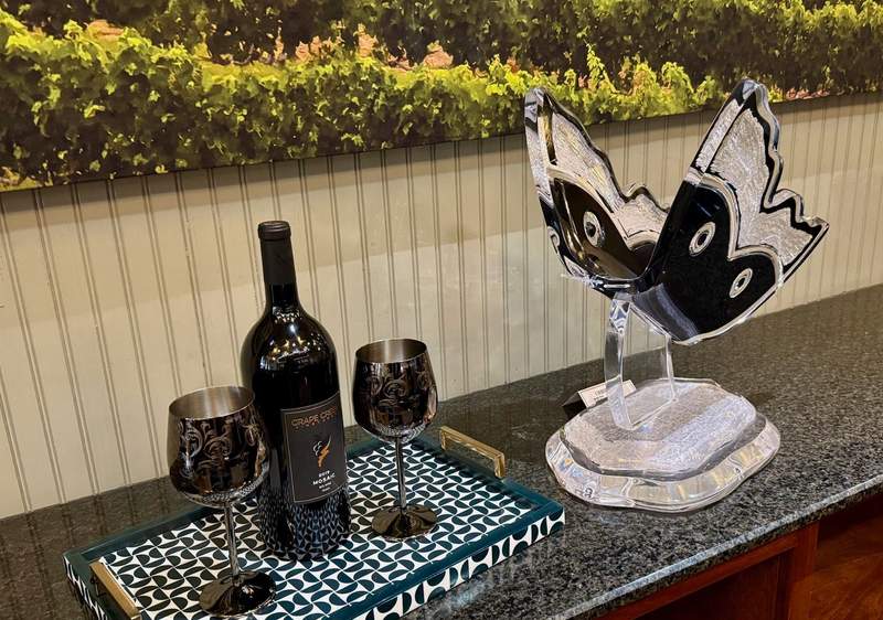 Black and white acryllic butterfly (or fairy?) watches over an open bottle of wine flanked by two long stemmed glasses on a granite ledge facing a dense garden of green hedges.