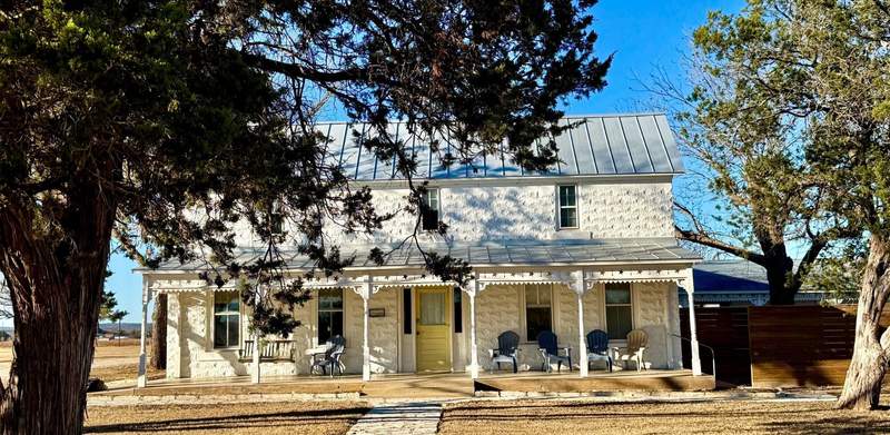 Cloudless blue sky frames a two story white house , with pecan tree taller than the roof on one side. Five chairs line the front poch where a wooden slatted swing to hold two adults hangs.