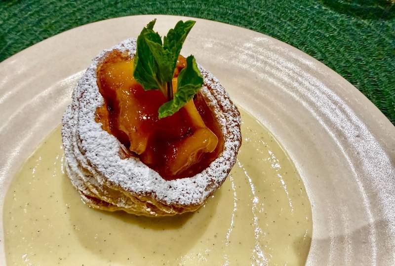 Peach segments poke out of a puff pastry dusted with powdered sugar. Three peppermint leaves stand tall in the peaches. The pastry on a round beige plate lathered in its center with luscious cream.