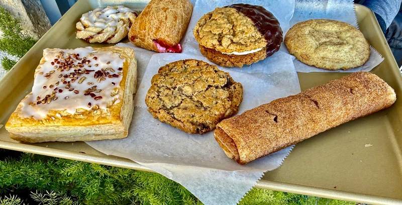 Seven large cookies and pastries fill a full-sized tray, resting on pieces of parchment paper. A bit of city greenery is visible.