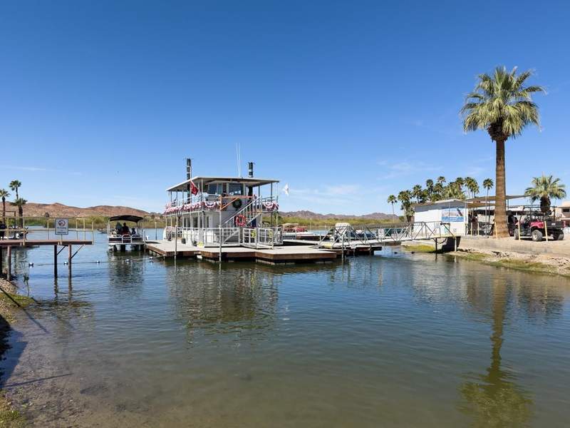 Paddlewheel boat docked at Martinez Lake in Yuma beneath a clear blue sky, with palm trees and calm water in view.