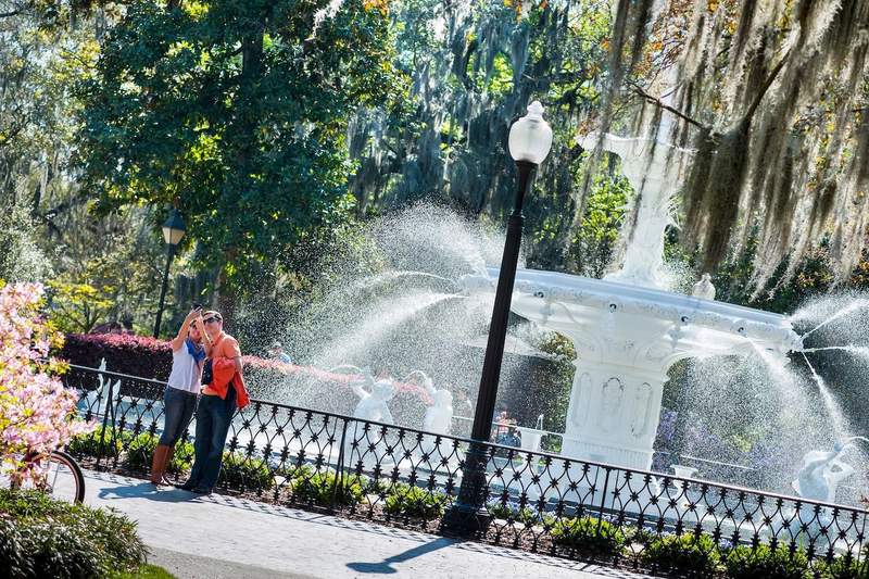 White Center Fountain surrounded by an iron fence, trees and flowers with couple taking selfie