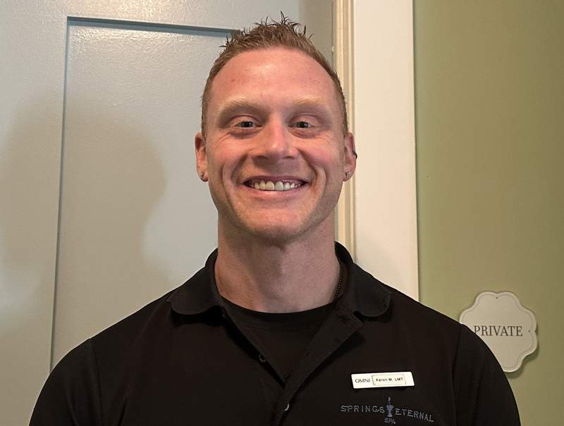 A smiling man with a dark shirt in front of a light wall.