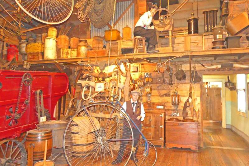 A period-dressed actor presenting a vintage penny-farthing high-wheel bicycle at the Oldest Store Museum exhibit.