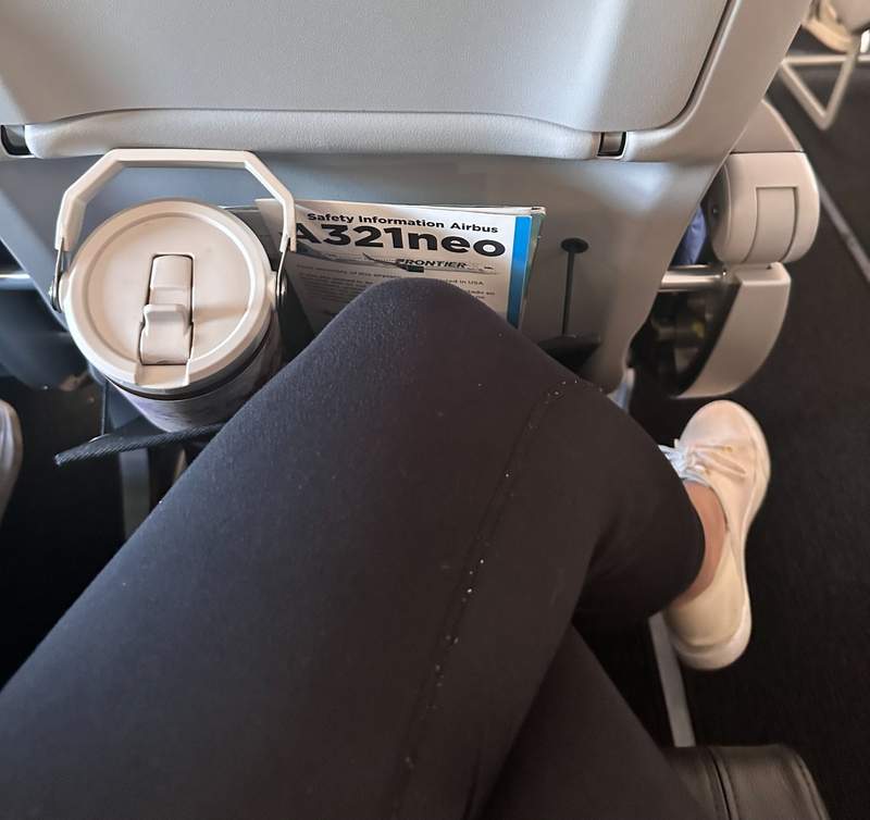 Knee with black legging and white shoe in an airplane seat with a water bottle in the seat back pocket.