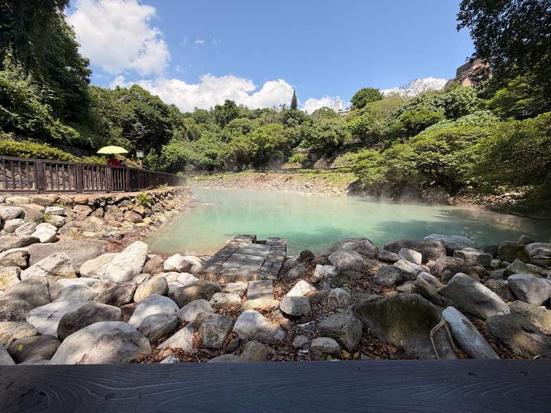 Large rocks surround a sea green smokey hot spring and a trail loops around the spring.