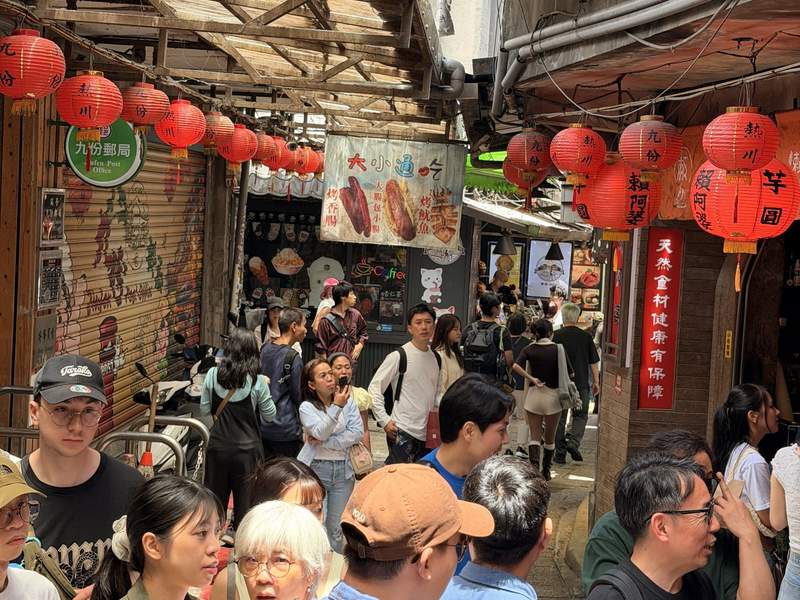 People strolling a narrow alley. Bright orange lanterns hang from the ceiling. 