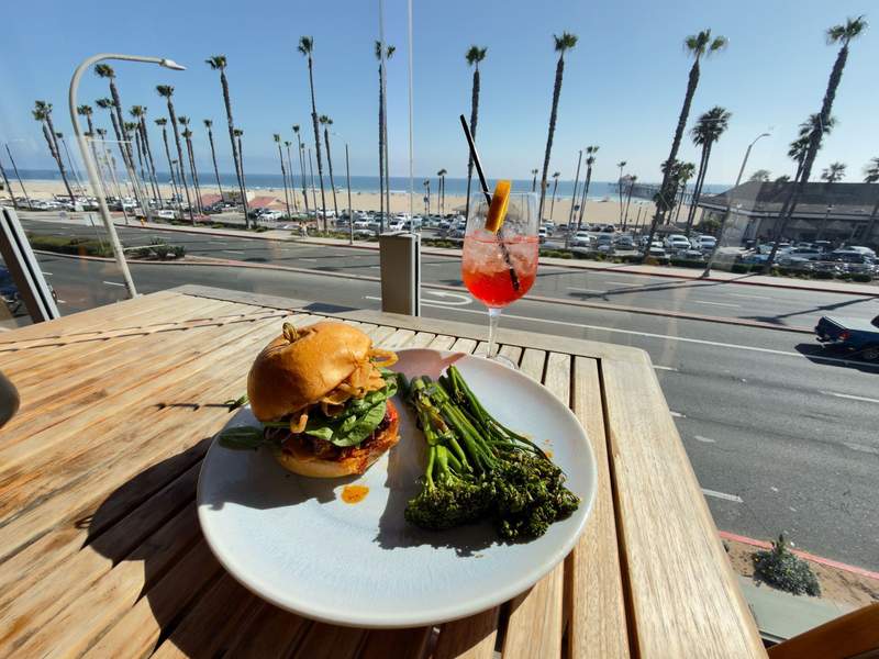 Fish sandwich with a side of broccolini and aperol spritz with huntington beach palm trees and ocean in the background