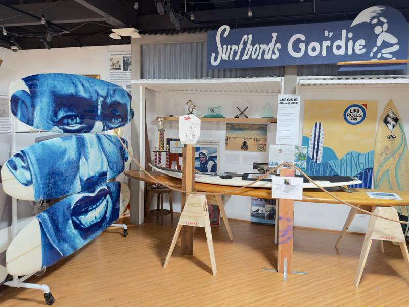 Surf legend Dukes face painted in blue over 3 surfboards. Other surfing inspired paintings and a sign hanging on the wall that says Surbords by Gordie.