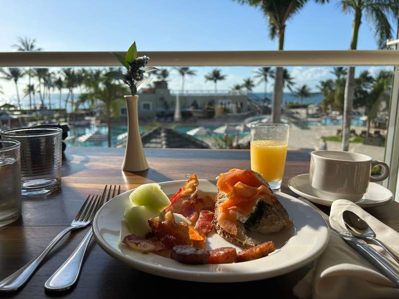 A hearty breakfast of toast and smoked salmon, fresh fruit, plantains, bacon and orange juice at the buffet restaurant with a view of the pool and beach in the background.