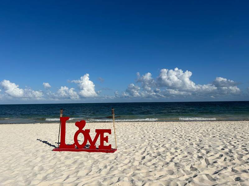 A large wooden LOVE sign in the sand set up by the hotel for romantic dinners. The ocean is in the background and the sky is bright blue with a few fluffy white clouds.
