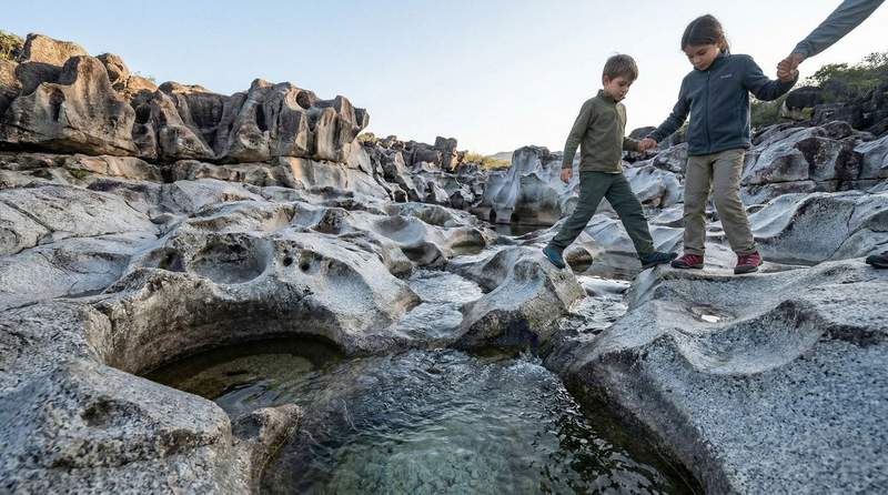 Detailed close-up of the grey granite rocks sculpted by the Sao Miguel River, with smooth circular formations and deep craters filled with crystal-clear water under the sunlight.