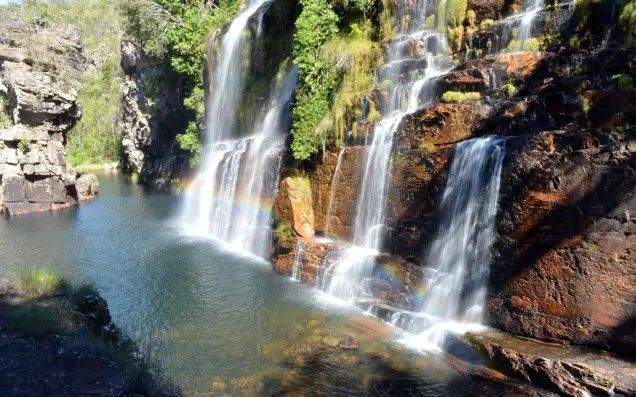 Waterfall cascades down a veil-like dark quartzite cliff face, falling into a deep pool surrounded by rocks and dense vegetation.