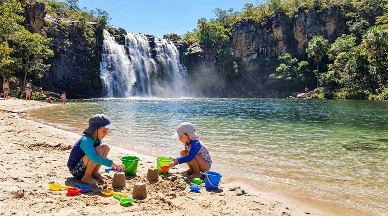 The beach at Poco Encantado, with children playing on sand at the edge of the emerald-green pool, with waterfall and dense vegetation in background.