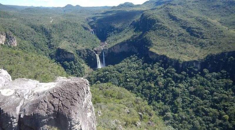 Chapada dos Veadeiros National Park. Above a deep river canyon, rocky cliffs with vegetation. In the distance, a waterfall plunges into valley, while mountains stretch toward horizon.