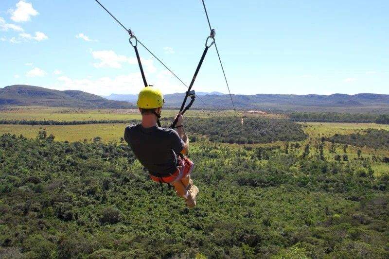 A person ziplining across a vast, lush valley with green mountain ranges in the background under a bright, sunny sky.
