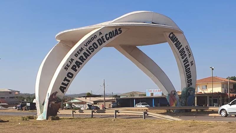 A large white concrete arch over a paved highway, with a welcome sign to Alto Paraiso de Goias, under a clear blue sky.