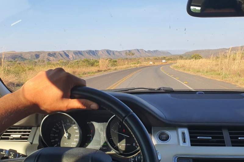 A first-person view from the driver's seat of a SUV on a paved road toward mountains of Chapada dos Veadeiros under a bright blue sky.