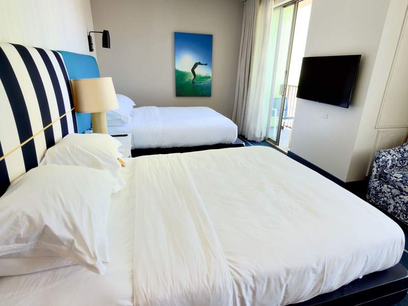 2 Queen bed room with striped headboard and white linens plus colorful image of surfer in a hotel room at the Kimpton Shorebreak Resort.