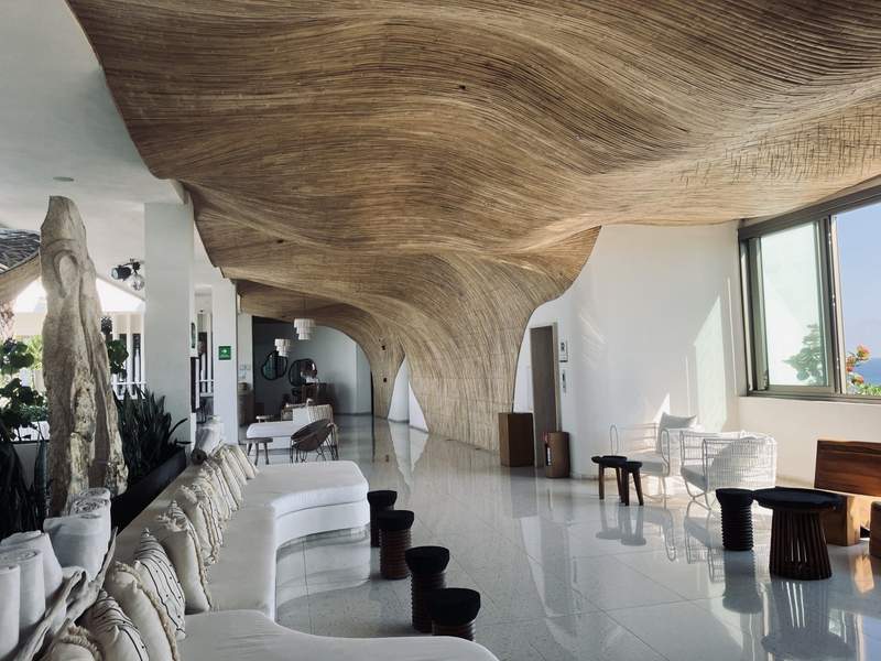 An intimate seating area for couples in an open-air hallway, with a long white couch, shining  white ceramic flooring and a natural wall feature reaching across the ceiling.