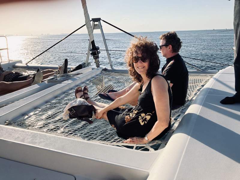 A smiling woman and man sitting at the bow of a catamaran on white netting, waiting for the sun to set.