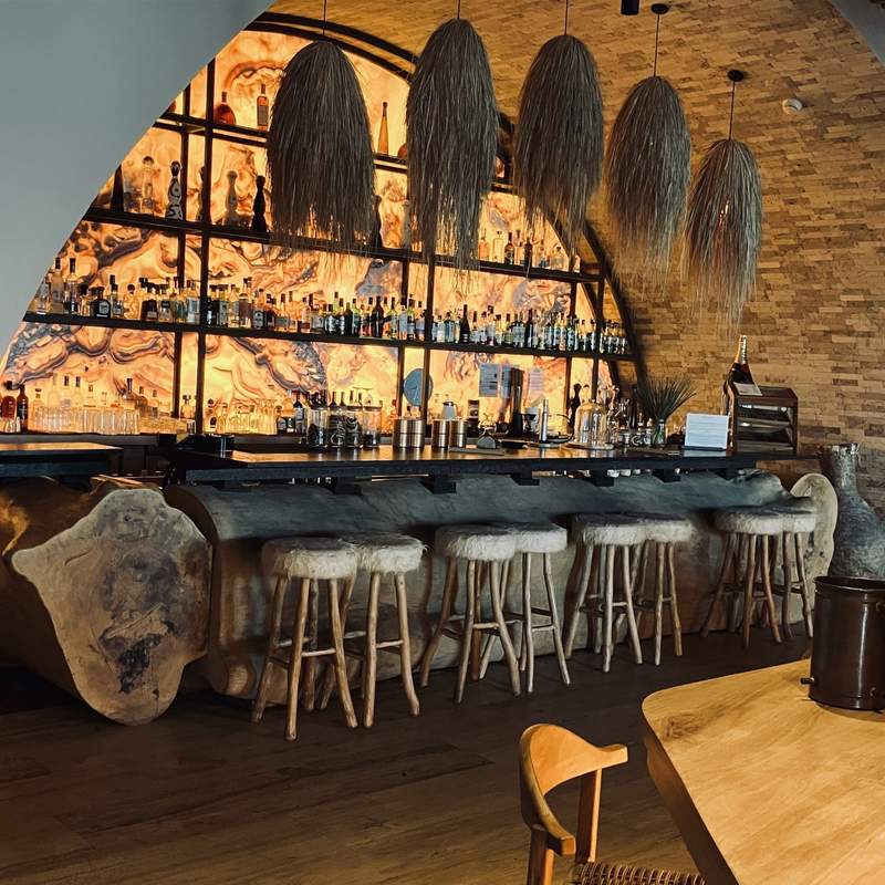 A dark brown bar with four high-stool sets for couples, with hanging light fixtures covered in long grasses, and backlit in a pattern of orange and brown.