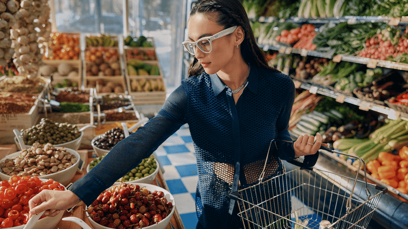 Brunette woman shopping in a produce aisle holding a wire basket and wearing Ray-Ban Meta AI Glasses.
