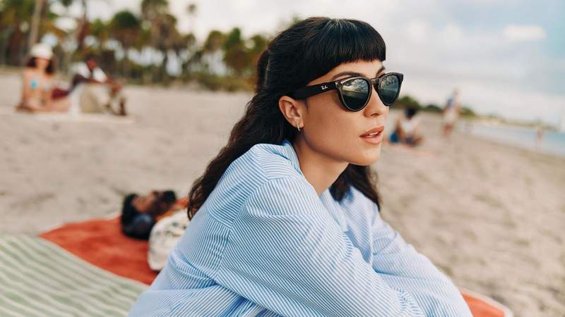 Brunette woman sitting on a beach wearing Ray-Ban Meta AI Glasses.