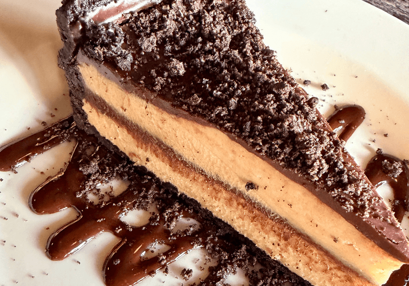 Slice of peanut butter chocolate cheesecake with Oreo crust and chocolate ganache drizzle on a white plate at Blueprint on 3rd in Birmingham, Alabama