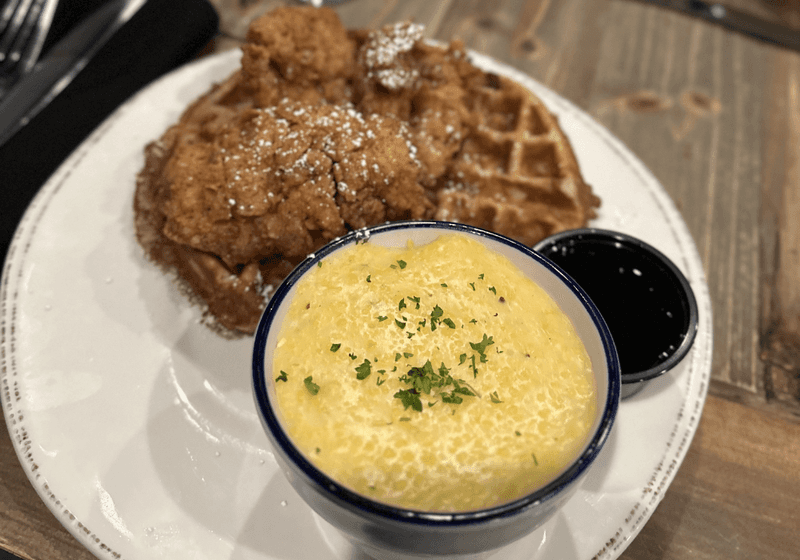Fried chicken and waffle with creamy cheese grits and syrup on a white plate at Farrelly's in Birmingham, Alabama