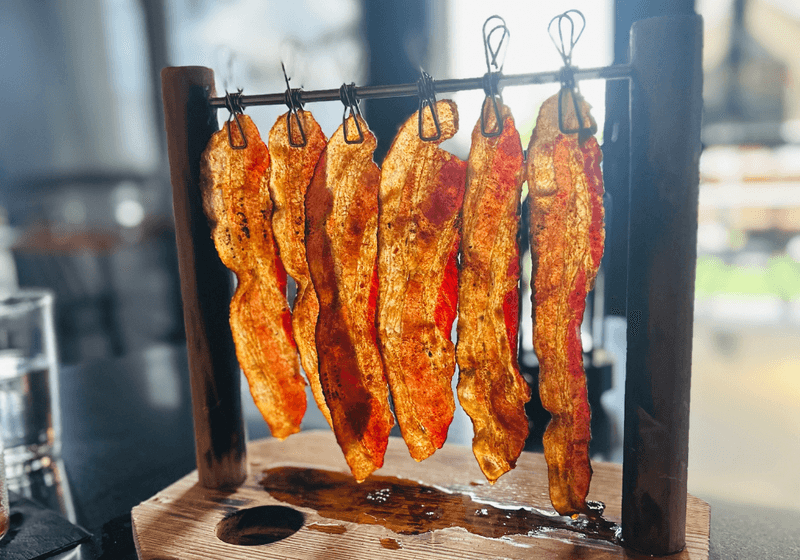 Five strips of crispy seasoned bacon hanging from a rustic wooden rack at Ironwood Kitchen and Cocktails in Birmingham, Alabama