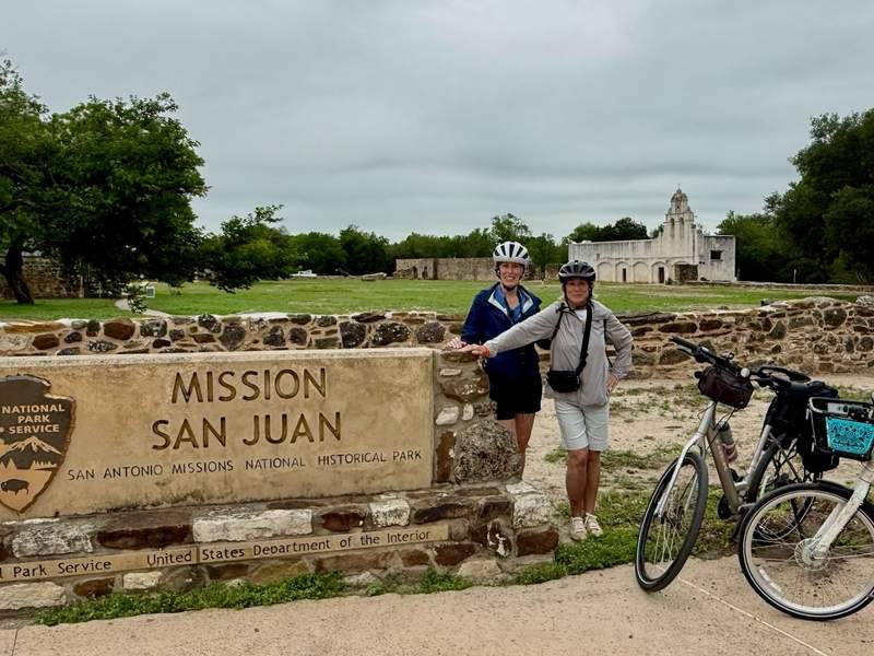 Two Women standing beside Mission San Juan sign with Mission San Juan Capistrano in the background.