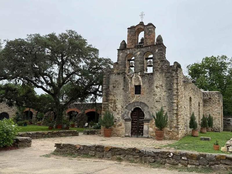 Stone mission with bell tower, large tree, and surrounding stone wall.