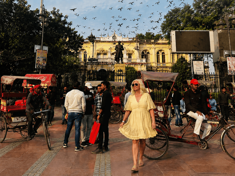 Woman walking through a busy Old Delhi square filled with rickshaws, people, and birds flying overhead in India.