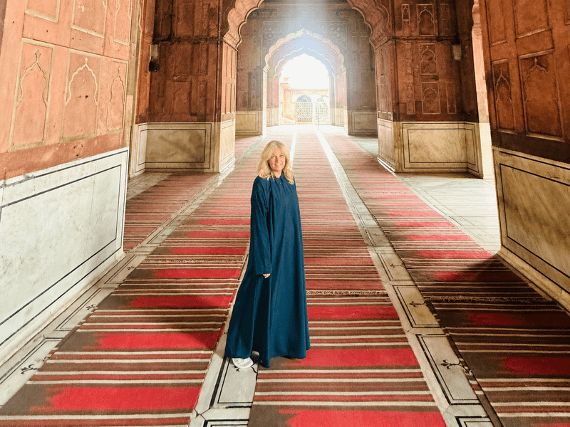 Woman standing inside jama masjid mosque in Delhi, India, surrounded by red carpets, arches, and warm sunlight streaming through the doorway.