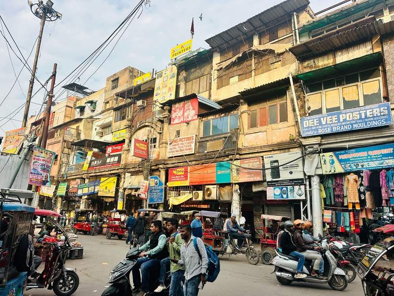 Busy street scene in Old Delhi with rickshaws, scooters, pedestrians, colorful storefronts, and aging buildings in India.