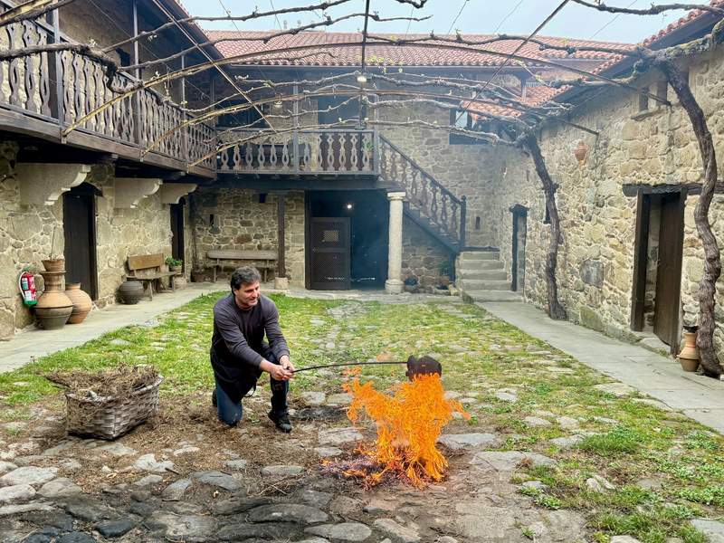 Man kneeling in Rectoral de Gundivós courtyard holding a ceramic pot over an open flame with a rod.